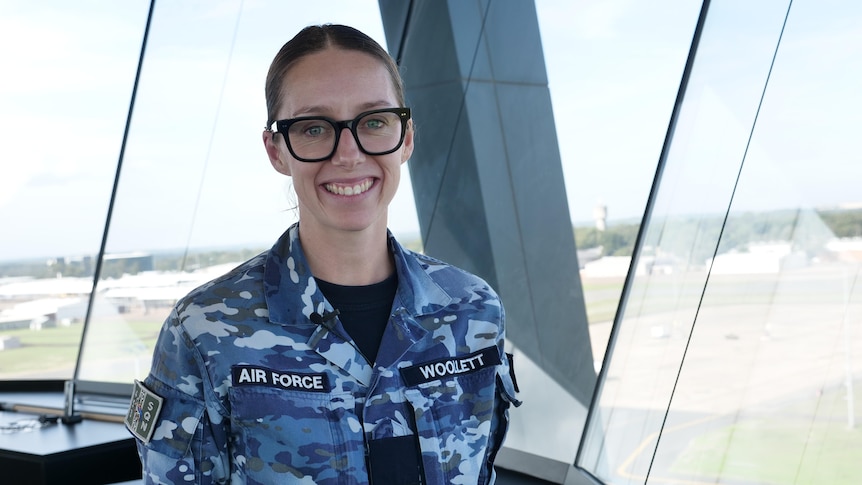 A smiling, bespectacled woman in an RAAF uniform stands in an air traffic control tower.