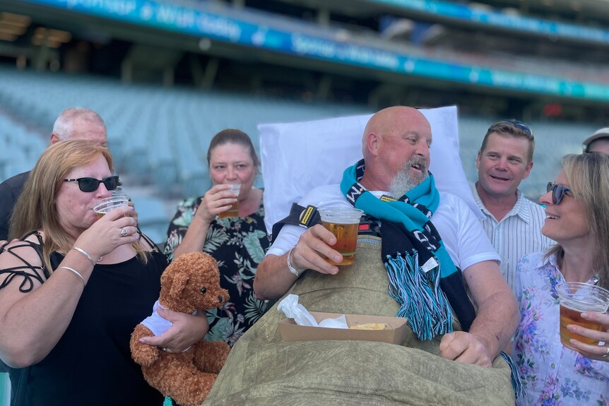A man lying on a stretcher having beer with his friends at an oval