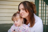  dark-haired woman sits on some steps, cuddling her young, fair-haired daughter.