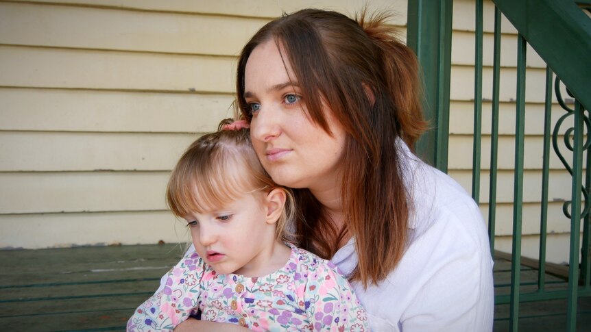  dark-haired woman sits on some steps, cuddling her young, fair-haired daughter.