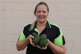A blonde woman holds a handful of avocados up to the camera and smiles.