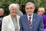 An older woman wearing a white jacket stands next to a man in a blue suit.