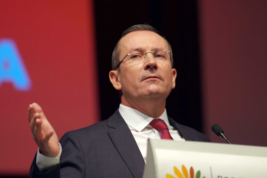 Mark McGowan standing behind a lectern with a stern look on his face.