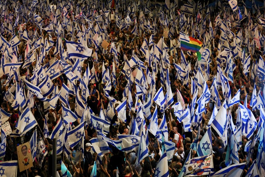 A large number of people holding Israeli flags at night, seen from a distance and an elevated position