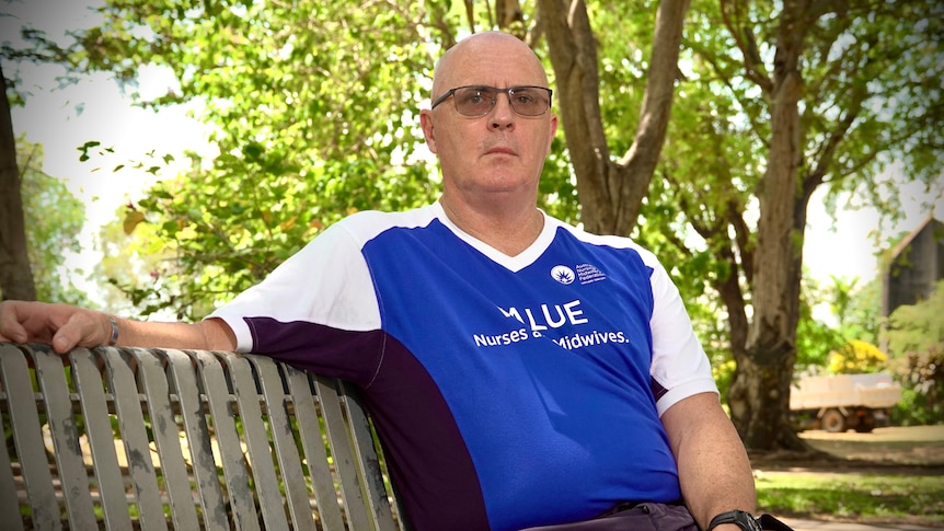 White man with bald head and glasses in blue shirt sits on park bench