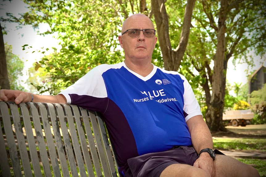White man with bald head and glasses in blue shirt sits on park bench
