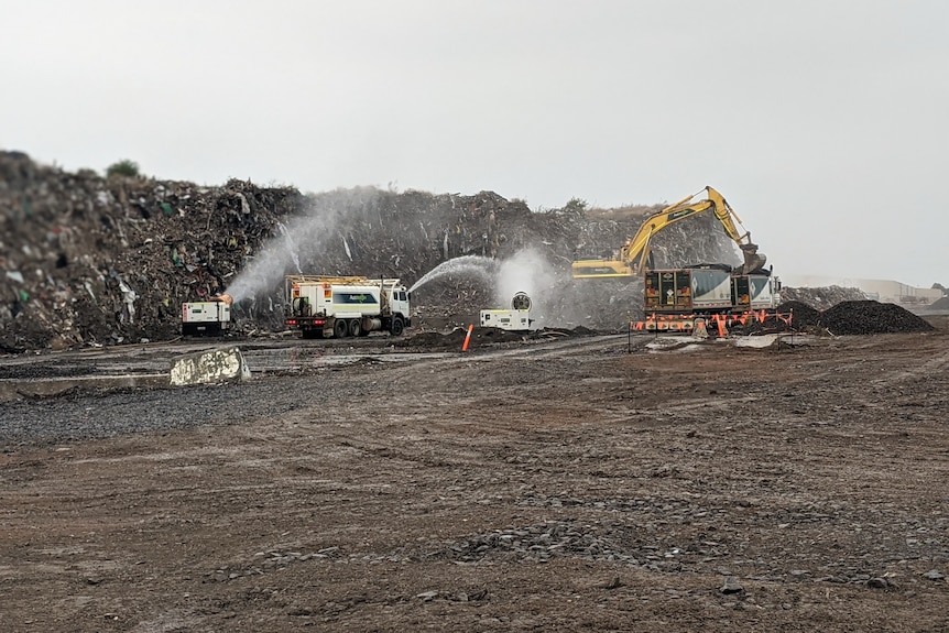 Trucks spray water on a large waste dump while diggers clear the waste into trucks. 