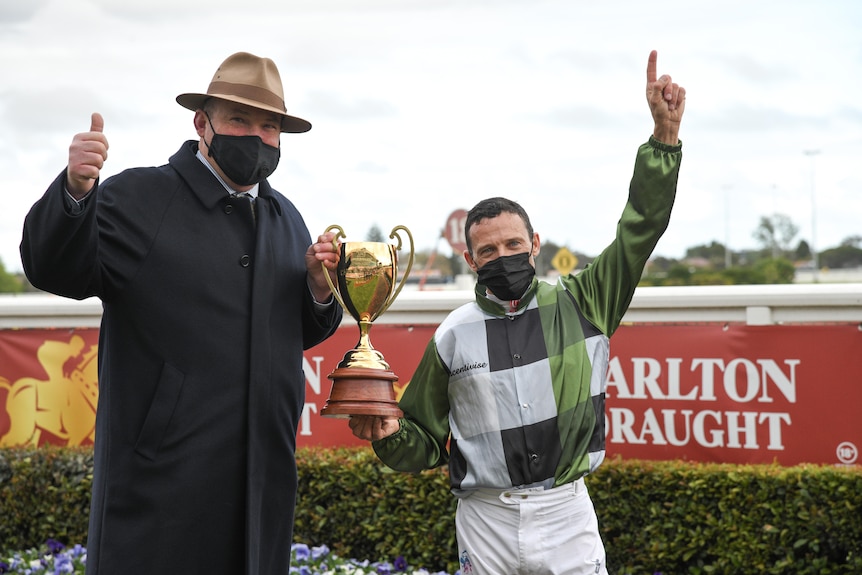 A jockey points to the sky and a coach gives a thumbs up while holding the Caulfield Cup trophy.