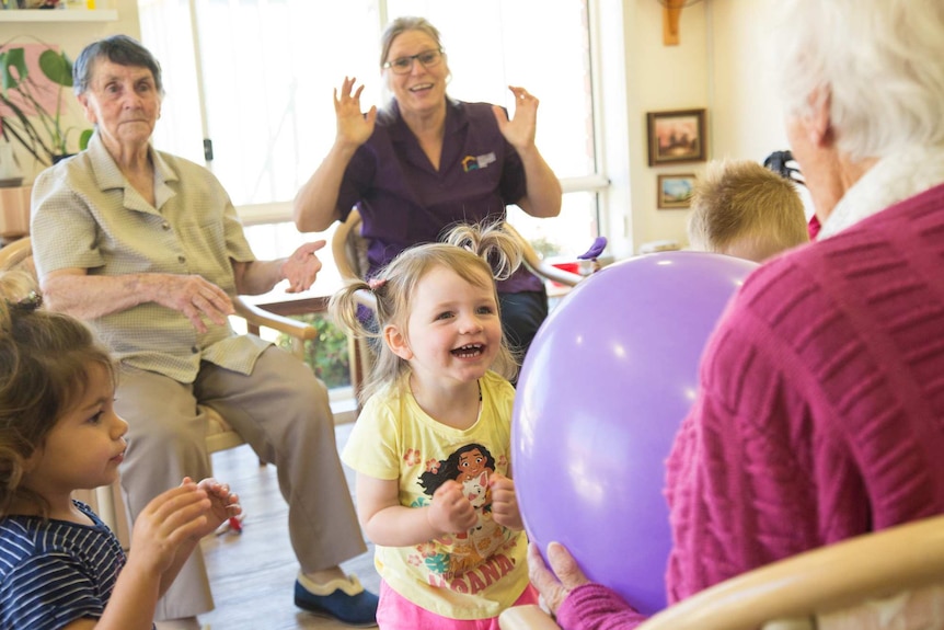 Elderly residents sit in a semi-circle throwing a large purple balloon around the room with three young children