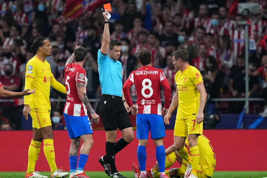 The referee holds up a red card as Antoine Griezmann looks on. A Liverpool player is sitting on the ground