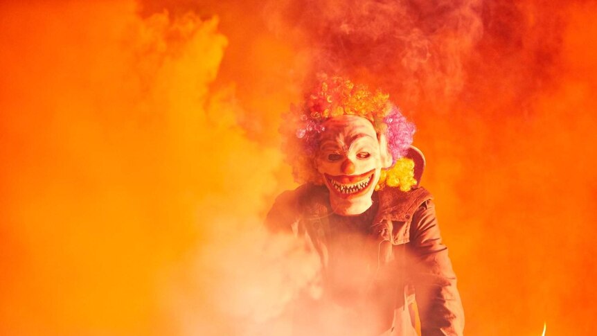A boy in a clown costume rides a BMX bike, surrounded by orange smoke.