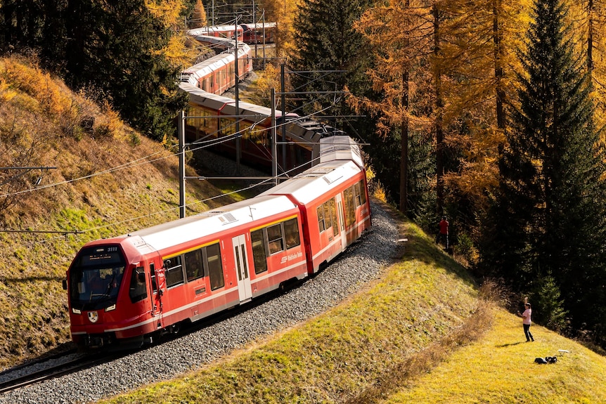 A red train on a winding track between a mountain and trees