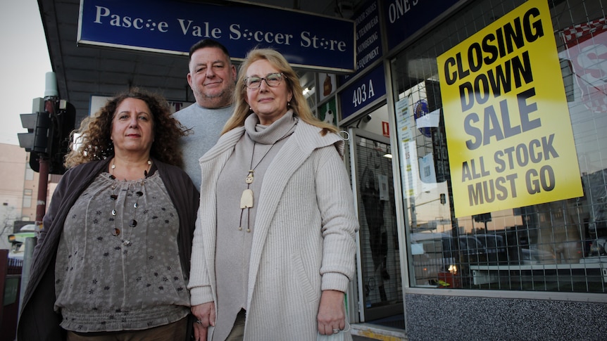 A man and two women stand in front of a Pascoe Vale Soccer Store sign. There is a yellow closing down sticker on the window