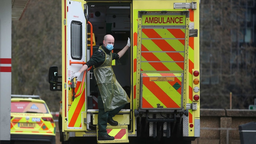 A man in a face mask and plastic apron stands in the doorway of an ambulance