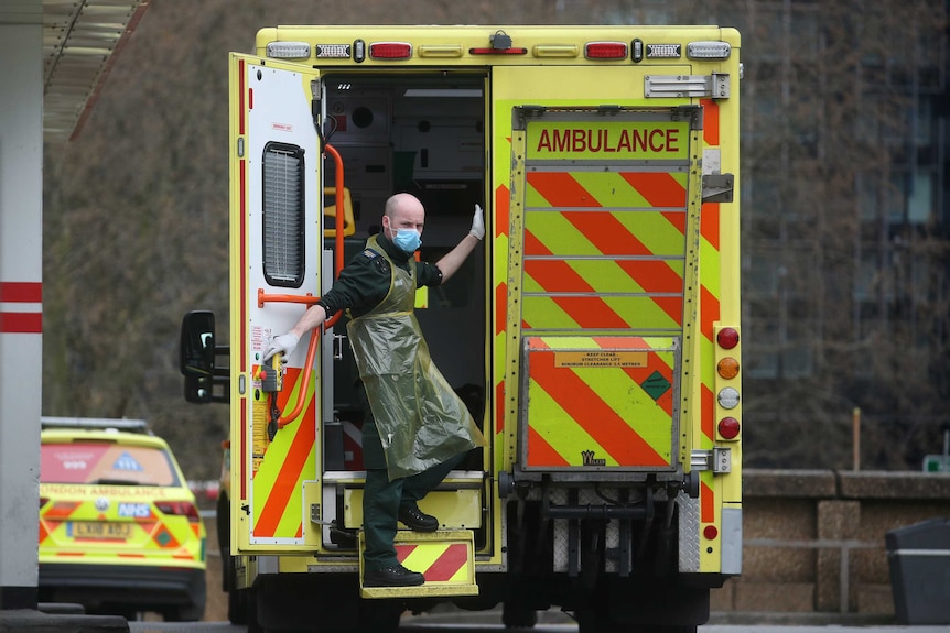 A man in a face mask and plastic apron stands in the doorway of an ambulance