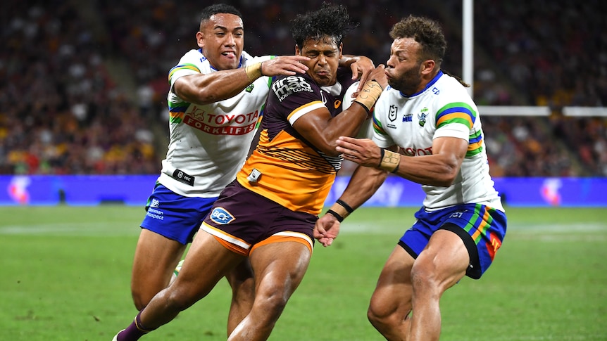 A Brisbane Broncos NRL player holds the ball as he is tackled by two Canberra Raiders opponents.