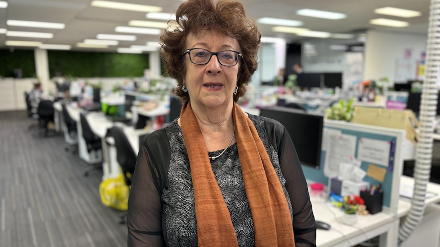 Woman wearing black top, orange scarf and black glasses looks at the camera. She is centre of frame, standing in an office.