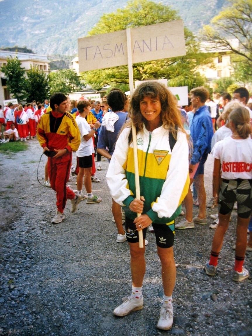 a woman with long hair is in Australian athletics clothes, holding a sign that says Tasmania