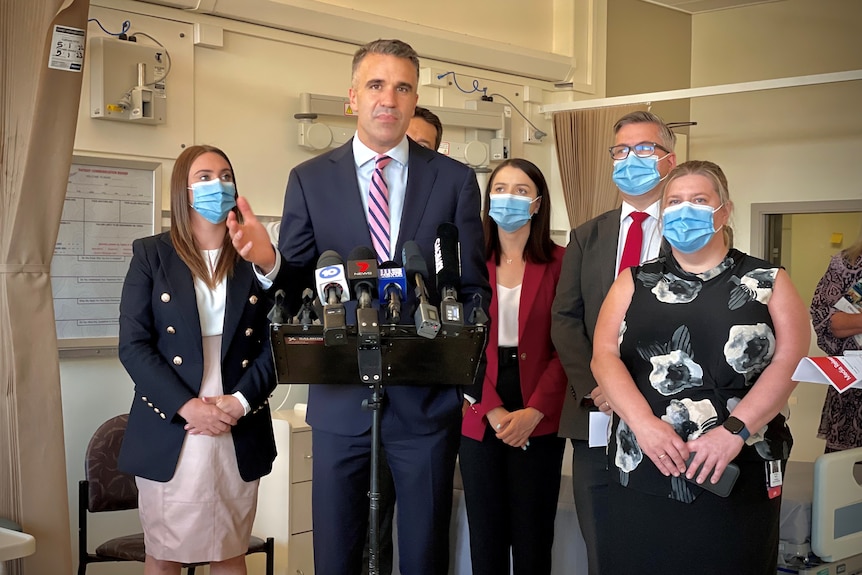 A group of politicians stand in a hospital room with media microphones in front of them