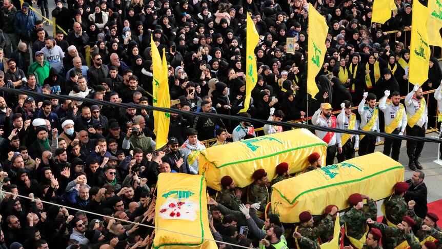 A large crown gathers around three caskets that are draped in yellow and green Hezbollah flag