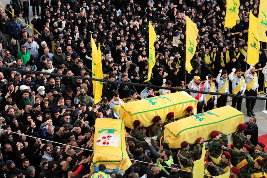 A large crown gathers around three caskets that are draped in yellow and green Hezbollah flag