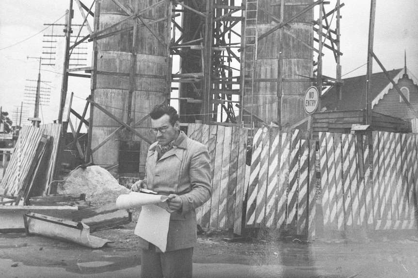 Old monochrome image of a man in smart jacket studying a plan in front of construction site.