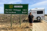 A man standing in front of a road sign and his caravan, smiling.
