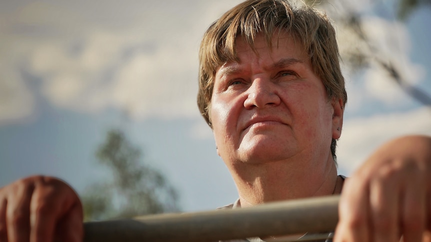 A woman stands with a fence in front of her with a neutral/serious expression.