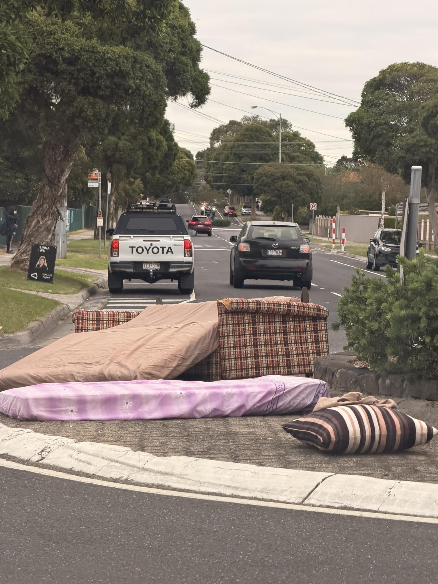 A couch and several mattresses and pillows, in the middle of a roundabout on a road. 