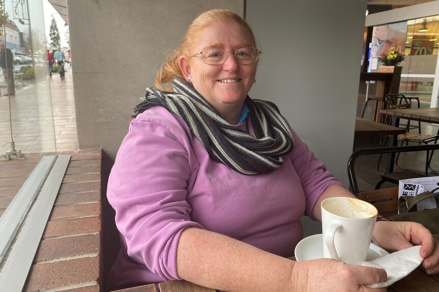 A woman with a scarf, wearing a purple jumper and glasses sits with a cup of coffee