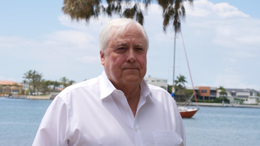 Clive Palmer with white hair wearing white shirt against blue water, sail boat and houses addressing media