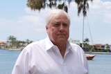 Clive Palmer with white hair wearing white shirt against blue water, sail boat and houses addressing media