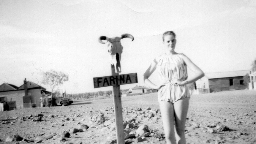 A young woman with a ponytail wearing a playsuit stands next to a stake in the ground that says 'Farina' with a ram skull on it.