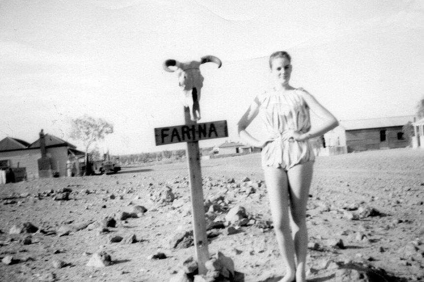 A young woman with a ponytail wearing a playsuit stands next to a stake in the ground that says 'Farina' with a ram skull on it.