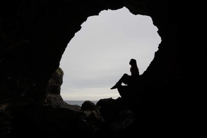 A silhouette of a woman sitting on a rock in a circular frame with the sea in the background.