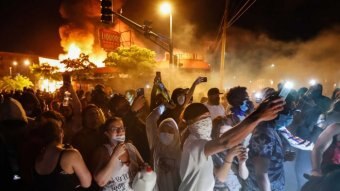 Hundreds of people stand in the street with a building behind them, with smoke billowing above it. Many film something not seen.