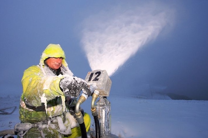 A man in yellow protective gear operates a snowmaking machine.