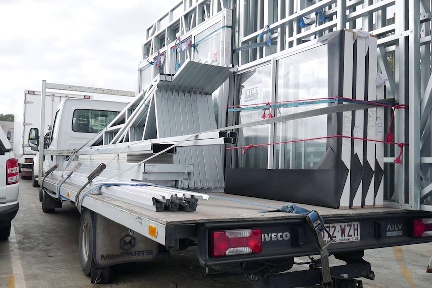 A utility vehicle carries building supplies alongside other vehicles in busy car park under clouds.