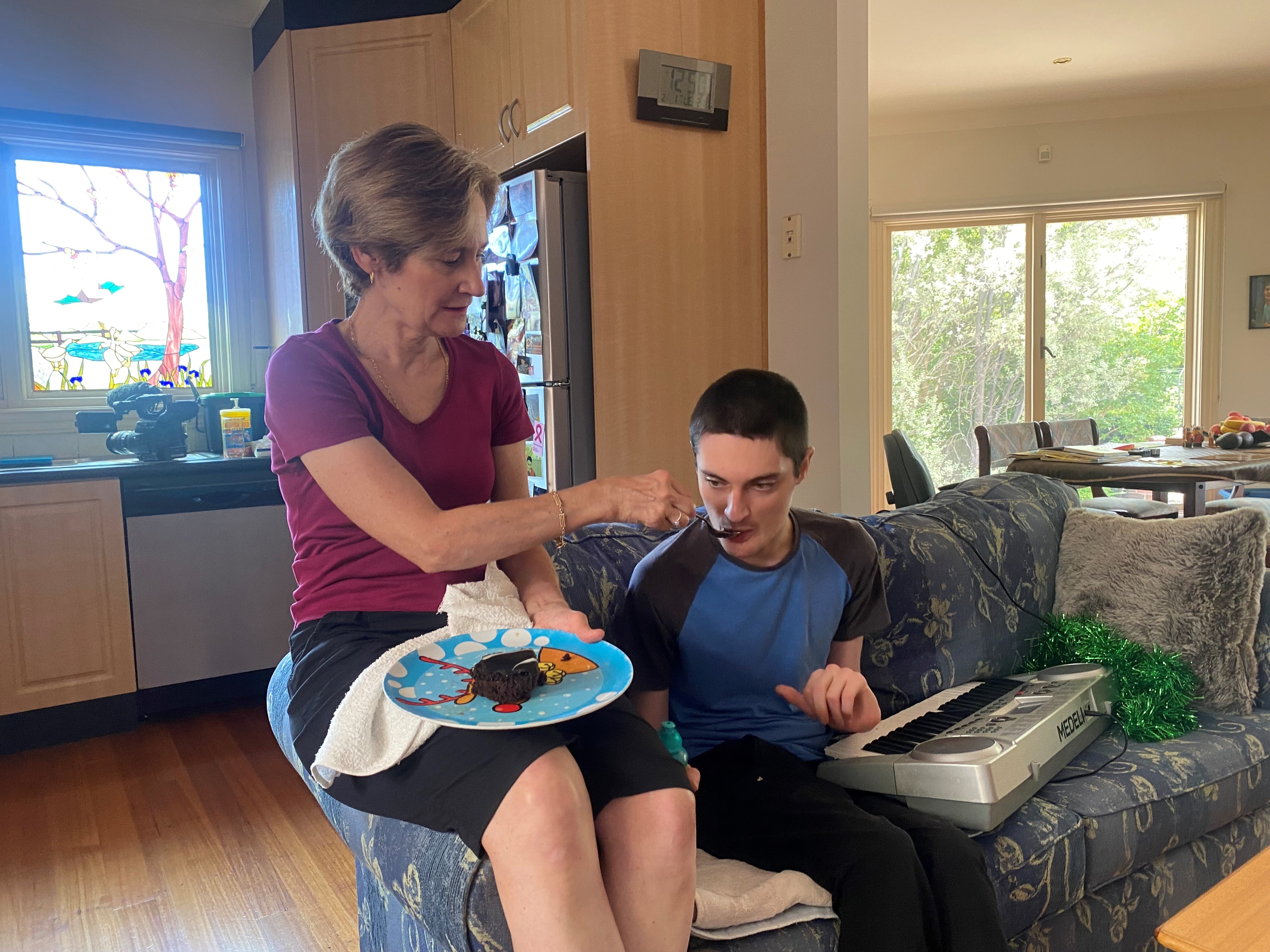 Karen McKenzie sits on the arm of a sofa and feeds her son Jarrod, who has an electric keyboard on his lap