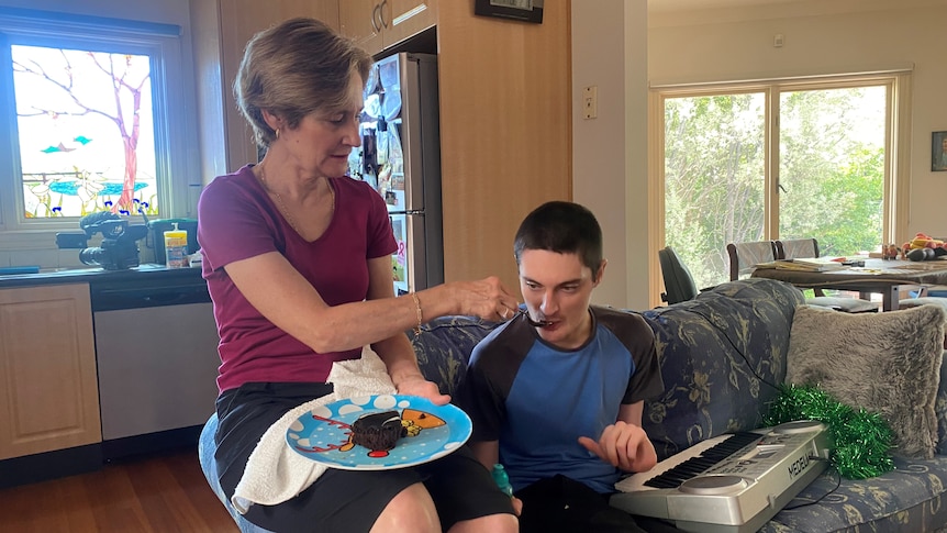 Karen McKenzie sits on the arm of a sofa and feeds her son Jarrod, who has an electric keyboard on his lap