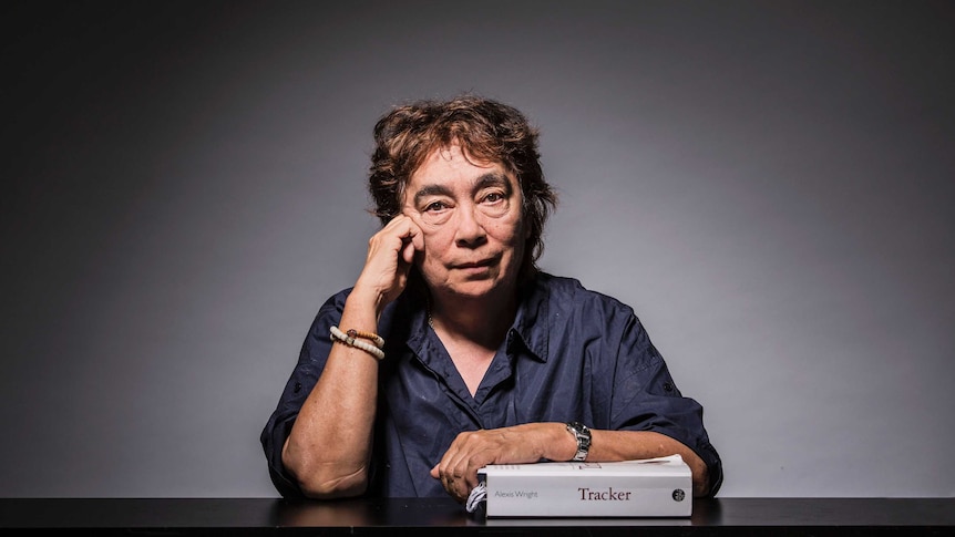 The writer sits, with cropped hair and wearing a dark blue shirt, with her arm leaning on a well-worn copy of her book Tracker.