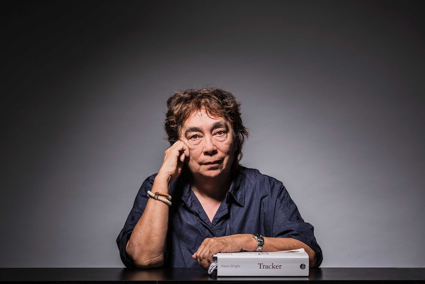 The writer sits, with cropped hair and wearing a dark blue shirt, with her arm leaning on a well-worn copy of her book Tracker.