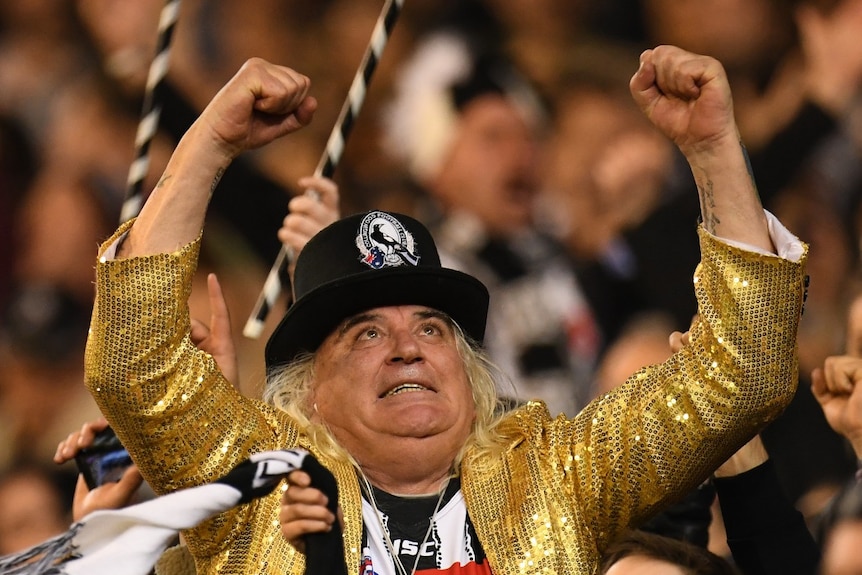 A man in a gold jacket and Collingwood hat celebrates in a crowd.