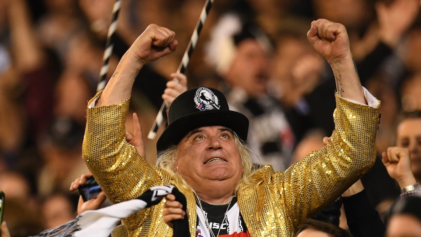 A man in a gold jacket and Collingwood hat celebrates in a crowd.
