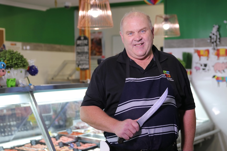 Paddy Sims stands in his butcher shop holding a large knife and wearing an apron