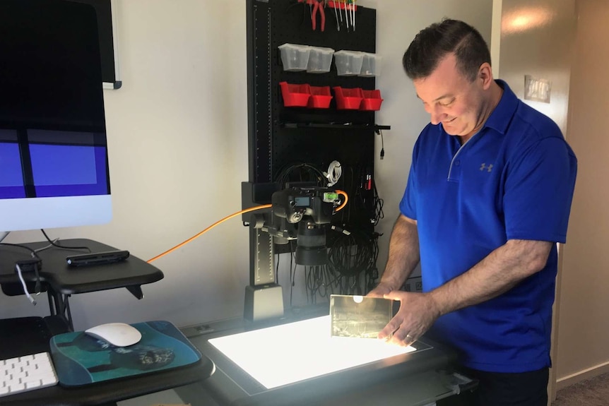 Photo of a man with black hair and blue tshirt, smiling, holding a glass negative, surrounded by photographic equipment