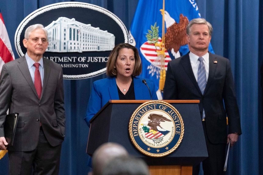 A female official and two male officials standing and speaking at a podium in front of US flag and a Department of Justice sign