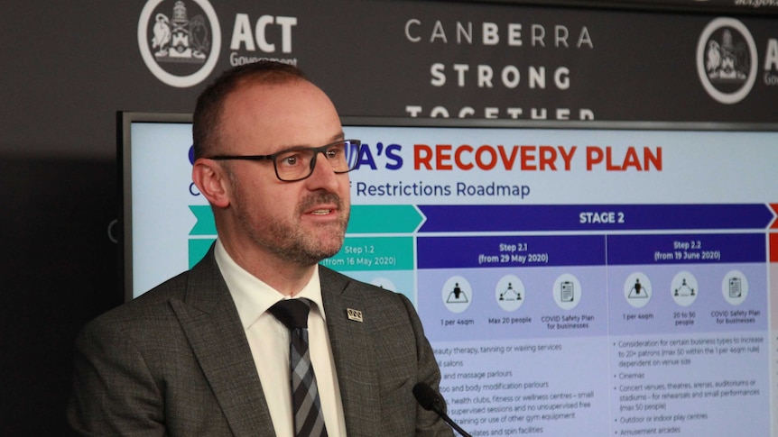 Andrew address the media, the recovery plan laid out on a screen behind him.