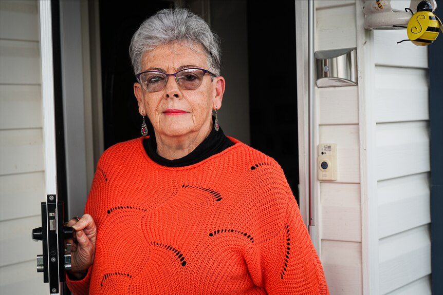 An older woman in a brightly-coloured jumper stands in the doorway of her home.