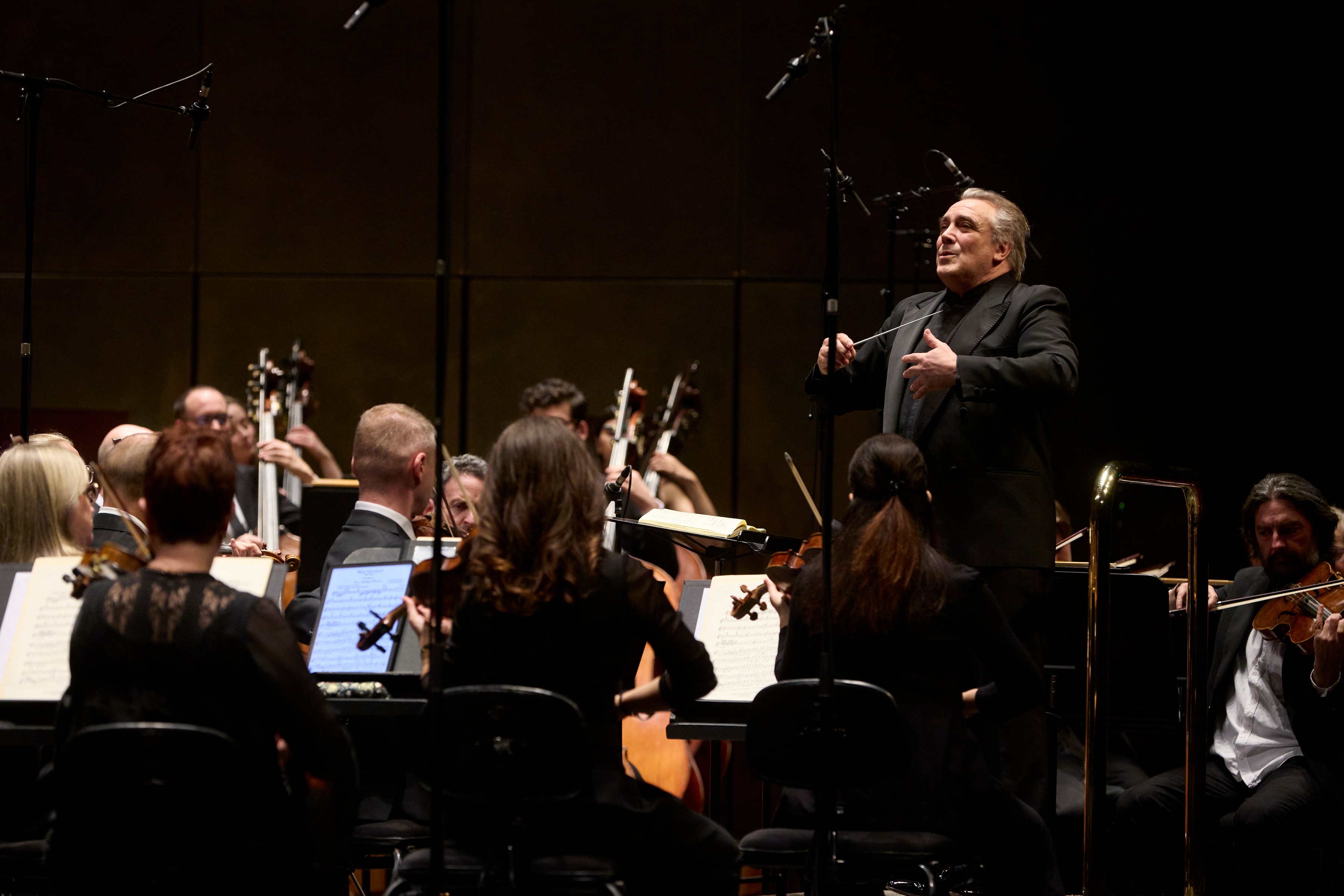 Jaime Martín conducts from a podium with a baton. He looks up and out at the orchestra as the musicians perform. 